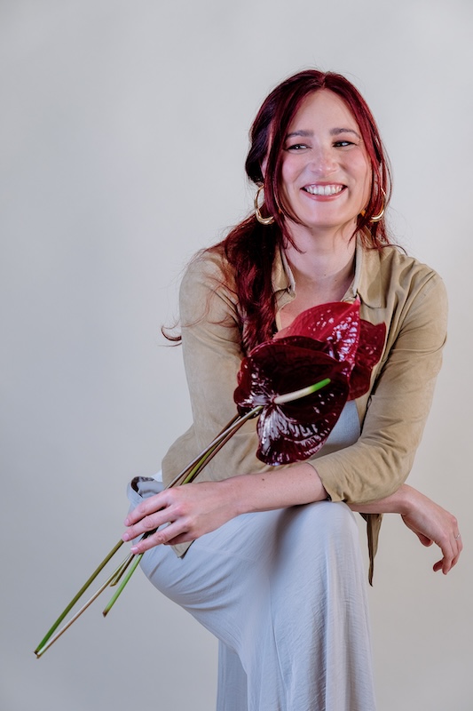 A woman with long red hair and gold hoop earrings smiles while sitting, embodying the elegance of Blooming Haus. She wears a beige jacket and a long light gray dress, holding a large dark red flower with glossy petals in hand. The background complements her look, being a plain light gray.