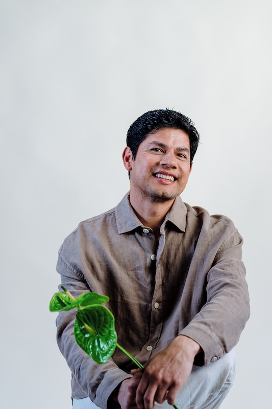 A man smiles warmly, seated against a plain white background. Clad in a light brown button-up shirt, his neatly styled short black hair complements his relaxed demeanor. In his right hand, he holds a green leafy plant from Blooming Haus, resting it gently on his knee.