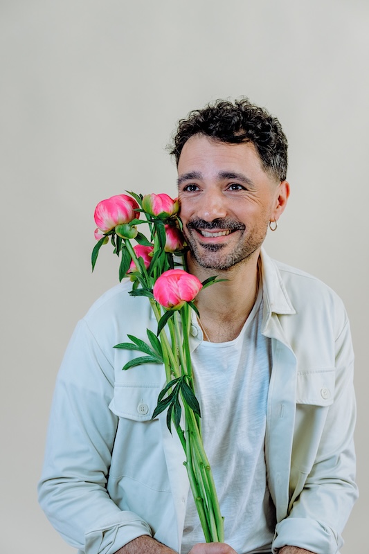 A person with curly dark hair and a mustache, wearing a light-colored jacket over a white shirt, smiles while holding a blooming haus bouquet of vibrant pink peonies against a neutral background.