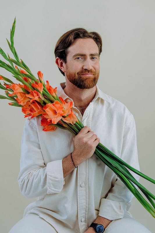A bearded man in a white shirt holds a bouquet of orange gladiolus flowers from Blooming Haus against a neutral background. He has light brown hair, a gentle smile, and wears a black watch on his left wrist. His right hand grips the flower stems, while his left rests on his lap.