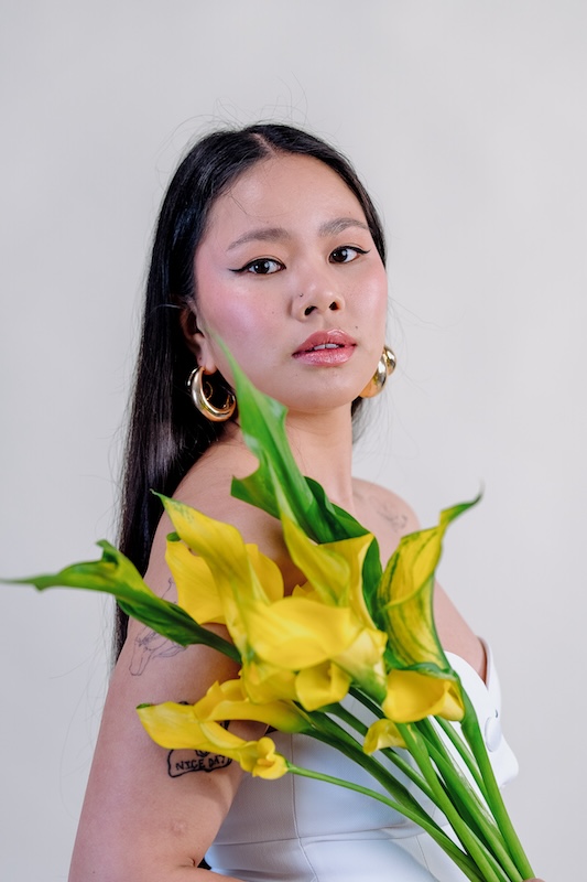 A woman with long dark hair and bold eyeliner holds a bouquet of yellow calla lilies from Blooming Haus. She's wearing large gold hoop earrings and a white strapless top. The background is a soft, neutral shade, enhancing the vibrant colors of the flowers and her subtle makeup.