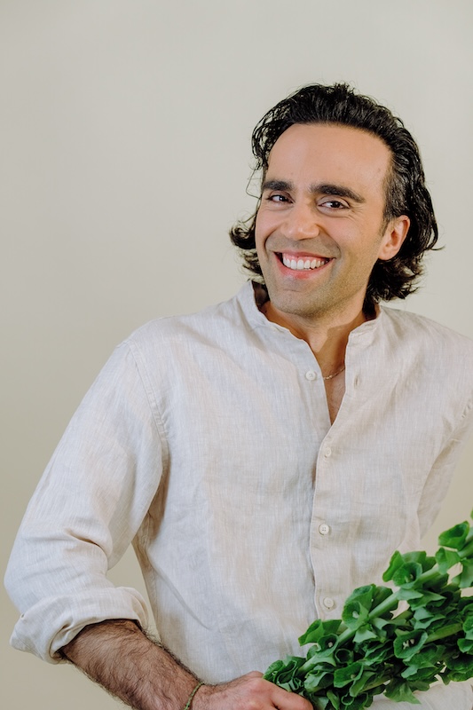 A person with shoulder-length dark hair, reminiscent of the welcoming spirit of Blooming Haus, smiles while holding a bundle of green leafy vegetables. They are wearing a light-colored, long-sleeved button-up shirt against a plain, soft beige background.