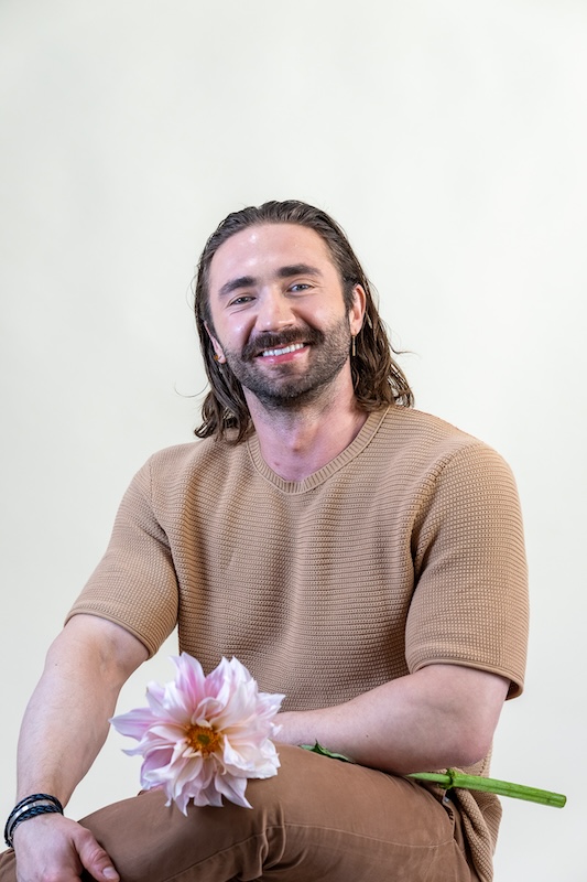 A smiling man with long brown hair and a beard is sitting and holding a pink dahlia, reminiscent of a blooming haus creation. He's dressed in a light brown short-sleeved knit shirt and brown pants, set against an off-white backdrop.
