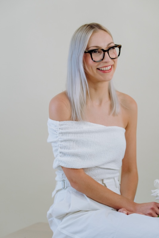 A woman with straight, light blonde hair and black-framed glasses smiles while seated. She wears an off-shoulder white textured top and high-waisted pants, exuding the elegance of blooming haus design. Her hands rest on her lap against a plain, light-colored background, creating a bright minimalist atmosphere.