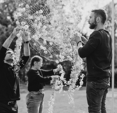 Three individuals work together outdoors to decorate a large net with flowers, reminiscent of an event handled by a London florist. The scene is in black and white, adding a timeless feel. One person stands on the right, elevating the net, while the others attach flowers below. They appear focused and collaborative, with trees visible in the background.