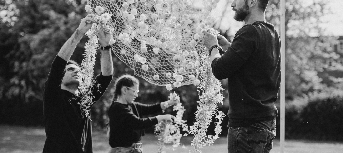 Three people are working together outdoors to hang decorative netting adorned with event flowers. The black-and-white photo captures two men and one woman focused on the task, each holding up a portion of the net. Trees and a lawn are visible in the background, creating a serene setting.