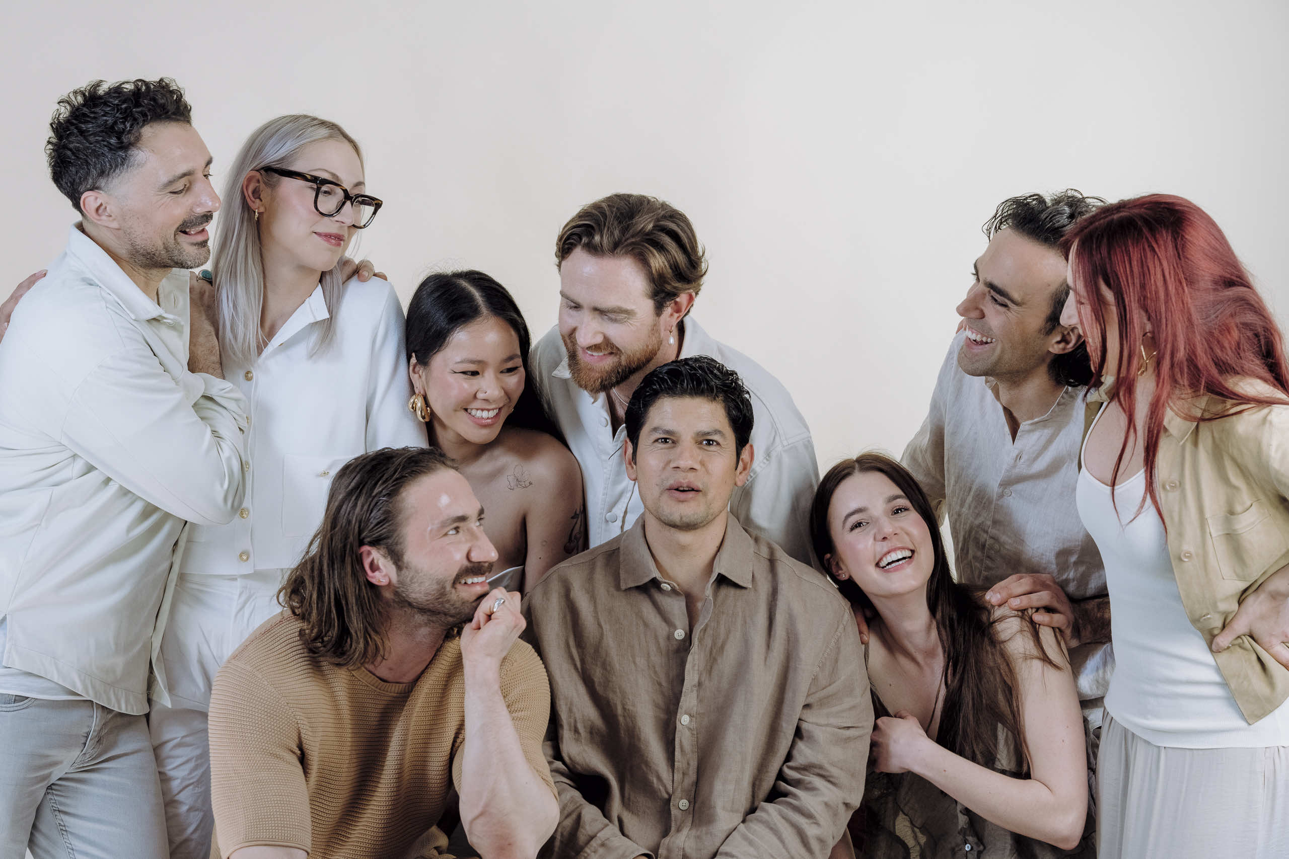 A group of nine Blooming Haus Team members stand closely together, smiling and interacting warmly. Casually dressed in neutral tones, they create a harmonious look. The plain off-white background keeps the focus on their happy expressions and relaxed poses.