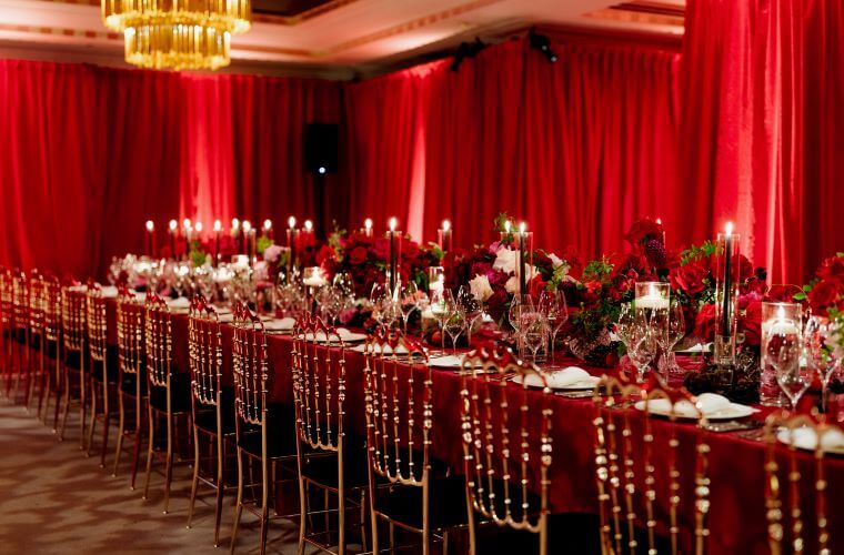 Long table set for an event with deep red flower centrepieces and a background of deep red curtains