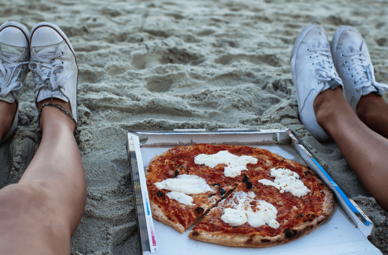 The feet of two people sitting on a beach with a pizza between them