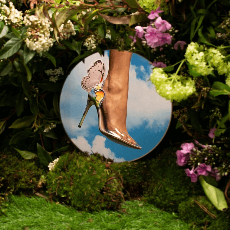 A shoe with a floral and butterfly design  in a circle against a backdrop of purple flowers and green foliage
