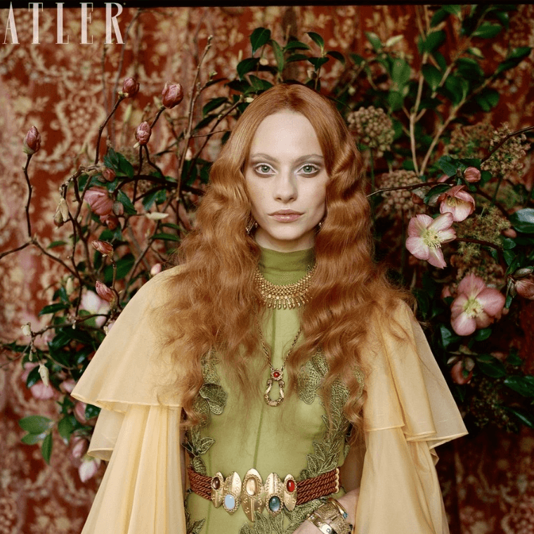 Woman with red hair dressed in rosetti era style fashion with blooming haus flowers as a backdrop