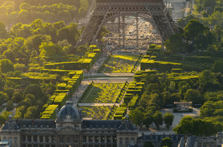 Aerial view of lush green gardens and tree-lined paths leading to the iconic eiffel tower in paris. People are scattered across the lawns, enjoying the sunny day. The foreground features a historic building with a domed roof, while olympic flowers bloom vibrantly, adding to the majestic backdrop of the tower.