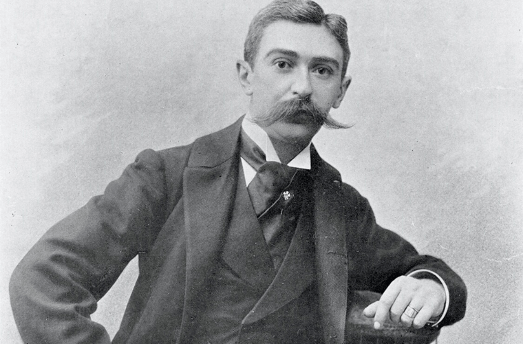 Black-and-white photo of baron baron pierre de coubertin of france with a thick mustache, wearing a three-piece suit, including a bow tie and a pocket watch chain. He has short hair and stares directly at the camera with a serious expression, leaning one arm against a surface adorned with olympic flowers while the other rests on his hip.