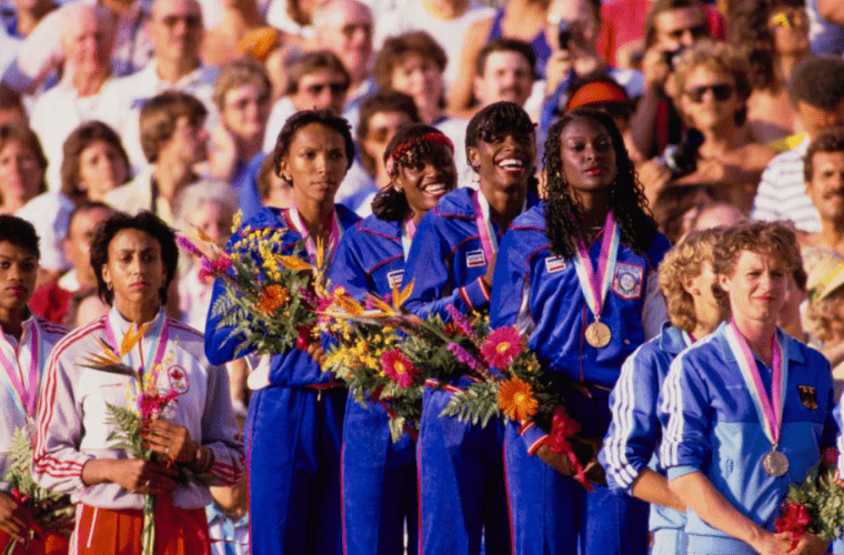 A group of six female athletes stand on a podium among a crowd of spectators. Five wear blue tracksuits and celebratory medals, while one wears a white and orange tracksuit. They all hold vibrant olympic flowers. The joyful crowd behind them appears to be celebrating the occasion with enthusiasm.