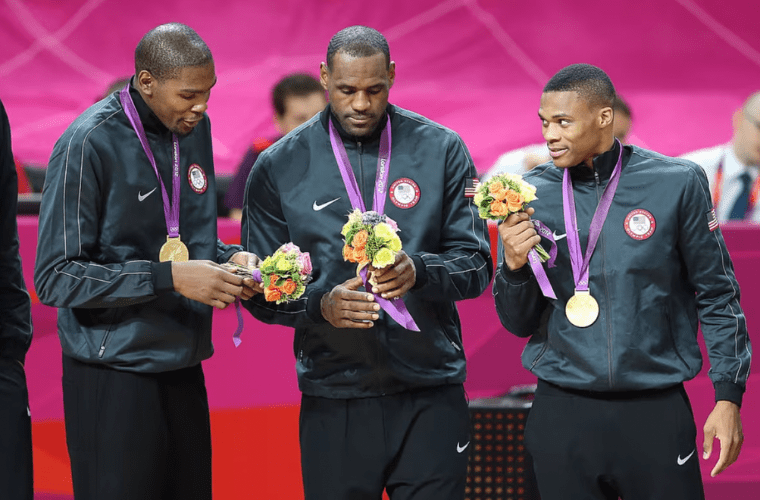 Three basketball players in team usa black jackets stand together, each wearing a gold medal. The player on the left holds an olympic flowers bouquet while looking down. The middle player, also holding a bouquet, gazes at his medal. The player on the right is smiling and glancing towards the middle player.