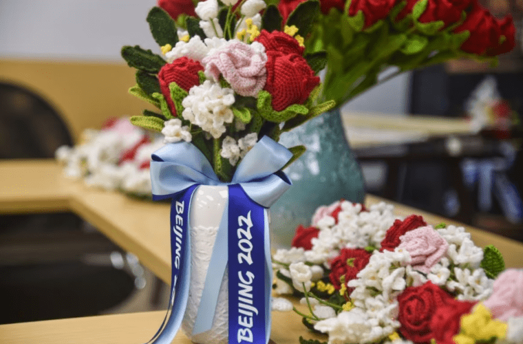 A bouquet of knit olympic flowers with red, pink, and white blooms and green leaves, arranged in a white vase with a blue ribbon labeled "beijing 2022. " the vase is on a wooden table surrounded by more knitted flower arrangements; a partially blurred background features chairs and a large green vase.