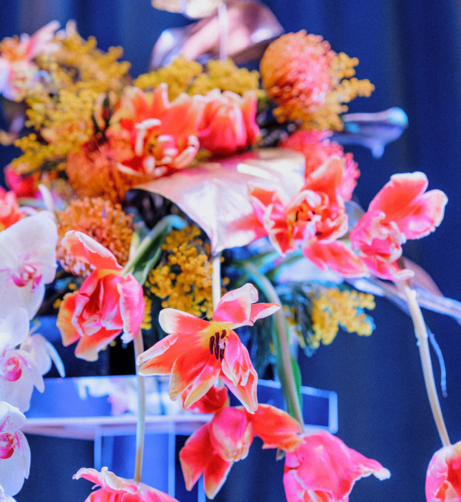 A vibrant floral arrangement by Blooming Haus features pink lilies, orange blooms, and white orchids interspersed with yellow sprigs. The flowers are displayed on a clear elevated stand against a dark blue background, creating a striking contrast.