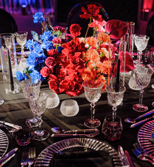 A lavishly set dining table boasts a striking floral centerpiece by Blooming Haus, showcasing red, blue, and orange blooms. Crystal glassware surrounds the arrangement, while small, textured candle holders sit on a black tablecloth, perfectly complemented by elegant plates and silverware.