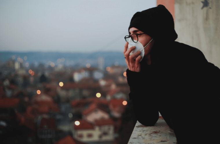 A man wearing a mask and looking out of a window to an urban street