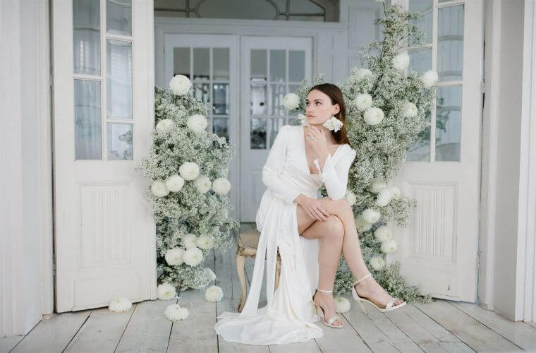 A bride seated in front of a white and green freestyle flower arch with the flowers coming half way up a doorway