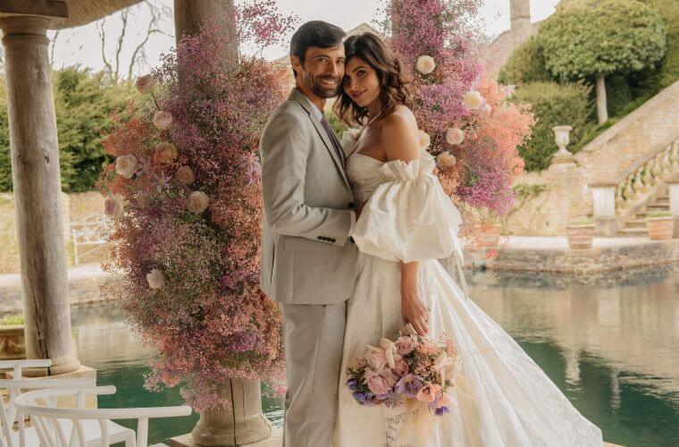 A wedding couple standing in front of a pastel coloured arch of flowers created by winding the flowers around pillars