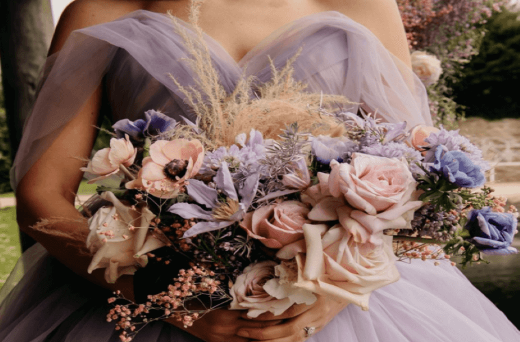 The torso of a bride wearing a lilac dress and holding a bouquet of lilac, pink, and peach flowers