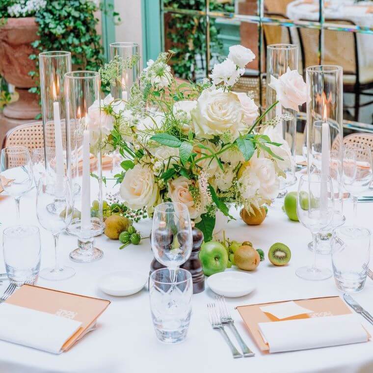 A wedding table set with a pale pink and white floral display, candles, fruit, and glasses