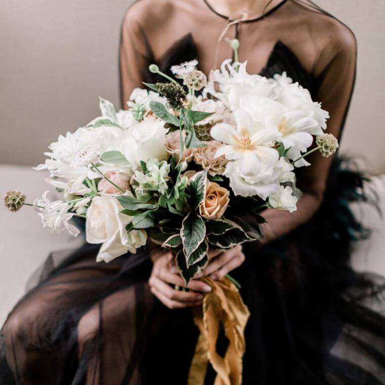 A bride in a black dress holding a bouquet of white and peach flowers tied with a gold ribbon