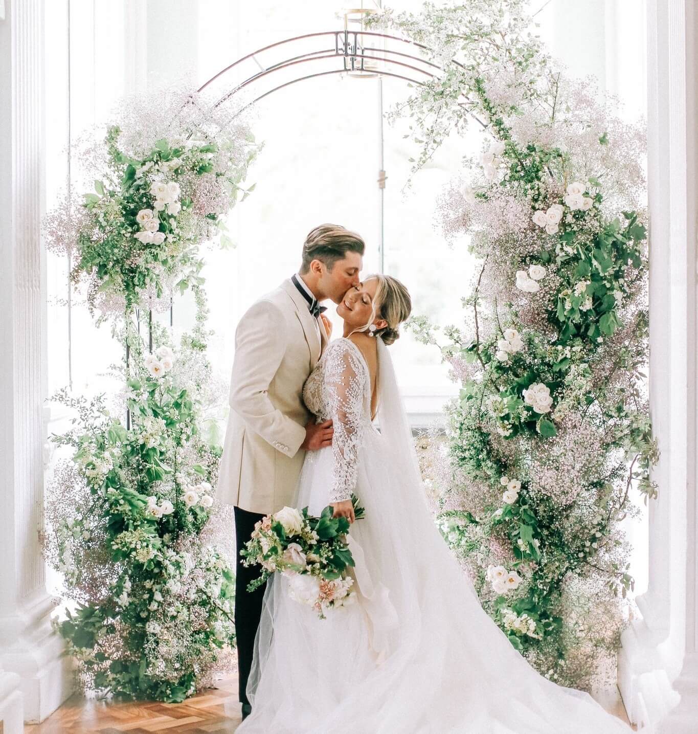 A couple standing beneath a wedding arch with white flowers and green foliage