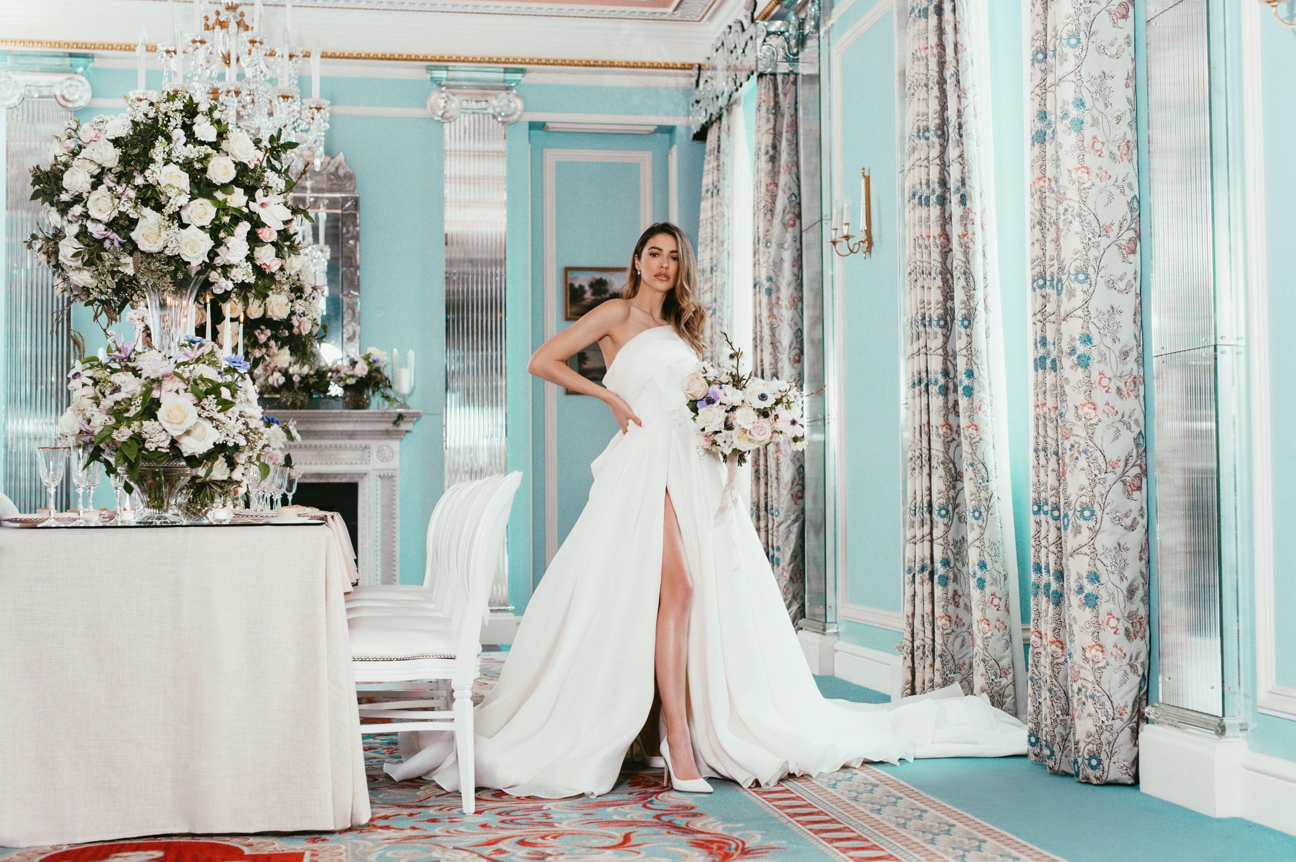 A bride in a white strapless gown with a thigh-high slit holds a bouquet from a renowned london florist and poses in an opulent room with turquoise walls, floral curtains, and elegant chandeliers. A dining table with white chairs and a tall, elaborate floral centerpiece is set nearby. The atmosphere exudes luxury and sophistication.