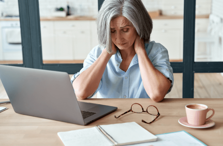 A woman sitting at a desk with a laptop in front of her holding her neck as though she is in pain